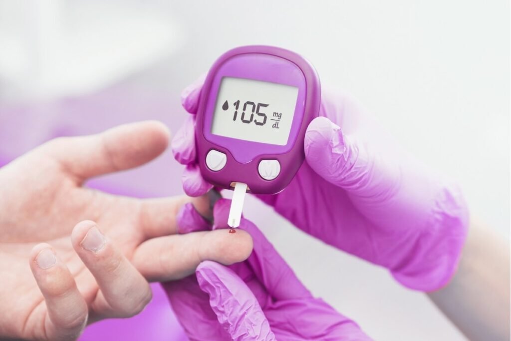 A close-up of a person with purple gloves checking blood glucose levels using a glucometer on a patient's finger. The screen shows a reading of 105 mg/dL.