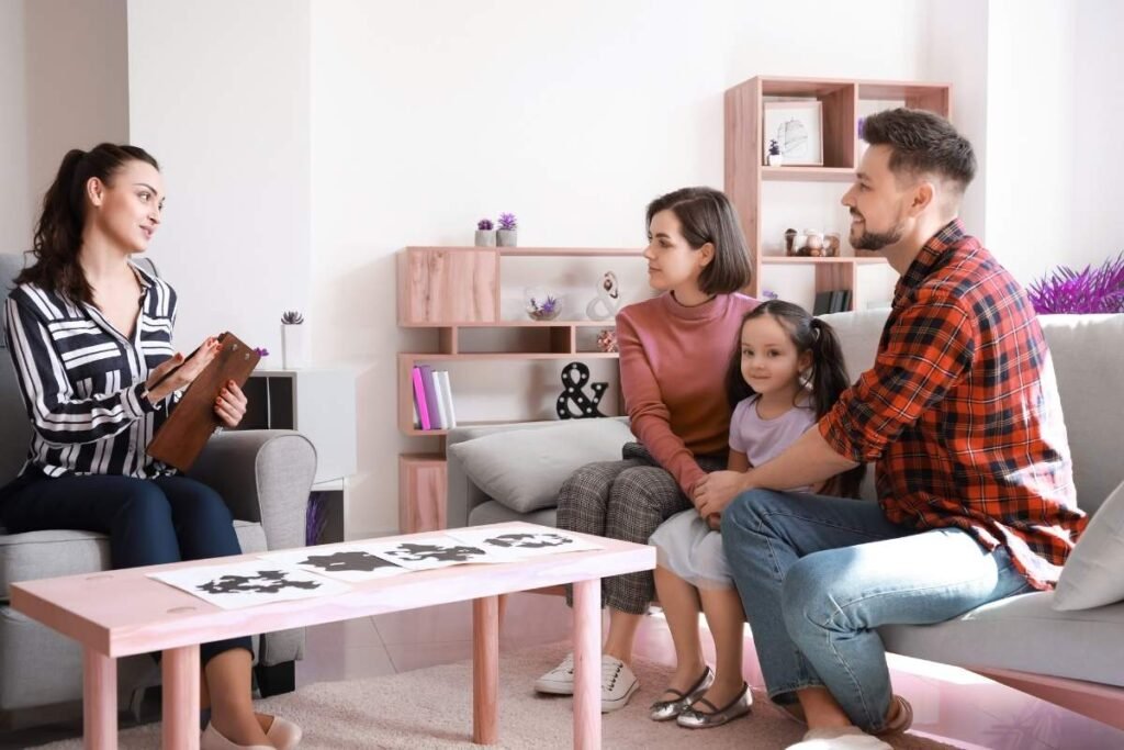 A therapist with a clipboard speaks to a family of three, consisting of a mother, father, and their young daughter, during a family therapy session in a cozy living room.