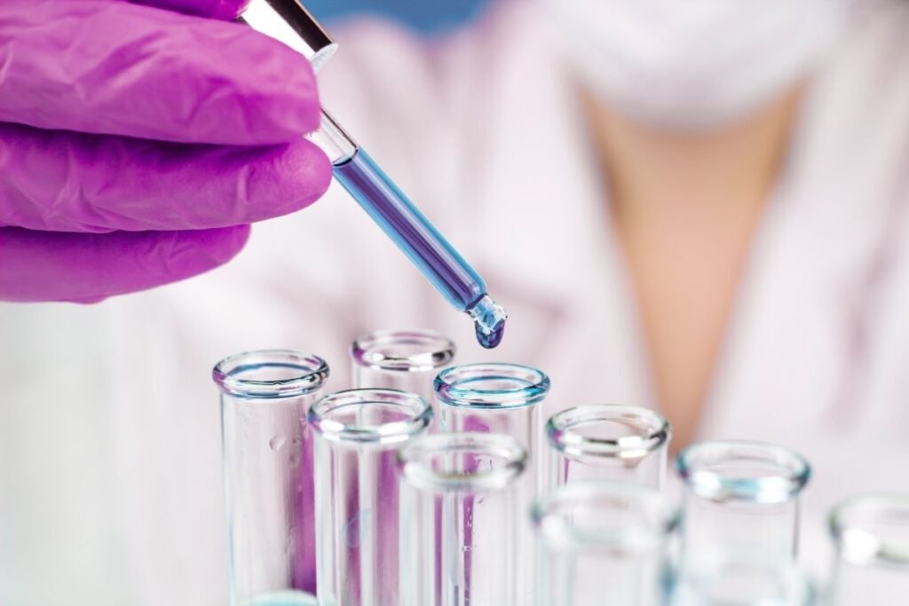 A laboratory technician in gloves using a dropper to add blue liquid to a test tube in a laboratory setting.