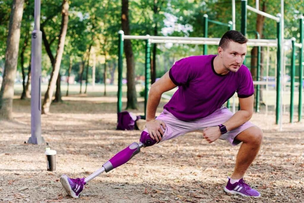 A man wearing a purple shirt and shorts exercises in a park using a prosthetic leg. He performs a deep squat with determination, showcasing personal strength and ability.