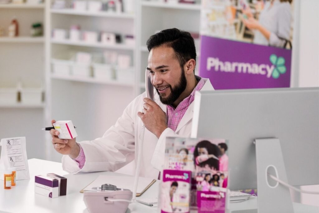A friendly pharmacist in a white lab coat talking on the phone while holding a medication box at a pharmacy counter.
