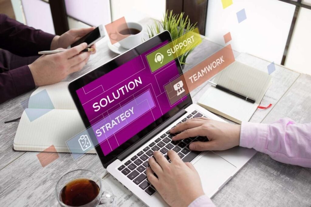 A person typing on a laptop displaying words like "Solution," "Strategy," "Support," and "Teamwork," with digital overlays, symbolizing smart solutions and strategies. A smartphone and a cup of coffee are visible in the background, indicating a collaborative work environment.